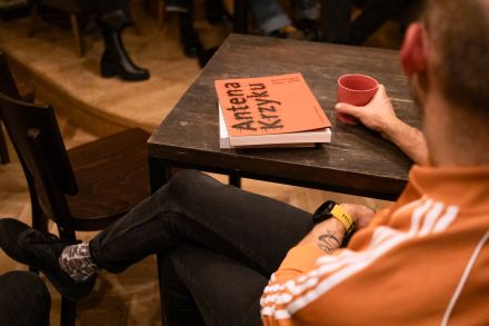 Przy stole siedzi mężczyzna. Trzyma w ręce szklankę z napojem i słucha dyskusji.