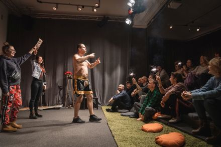 Na środku sali stoi artysta. Za mim dwie dziewczyny z widowni stoją z uniesionymi rękami. Publiczność się śmieje.