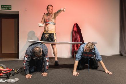 Dwaj mężczyźni są na czworaka na podłodze. Artysta kładzie im na plecy świnię z pleksy.