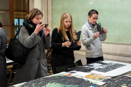 Kobieta i mężczyzna oglądają wycinanki, biorą je do rąk. Inna kobieta robi zdjęcie pracom.
