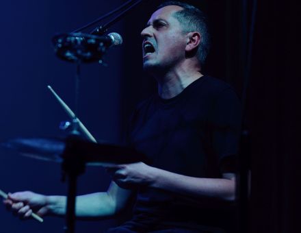 Zbliżenie na twarz muzyka. To wokalista, który gra na perkusji.