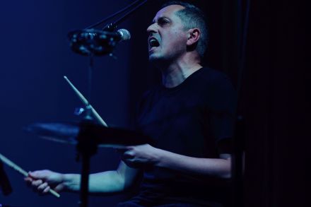 Zbliżenie na twarz muzyka. To wokalista, który gra na perkusji.