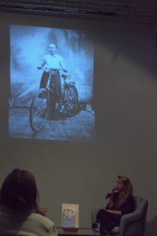 Na  ścianie wyświetlono slajd z kobietą trzymającą kierownicę roweru, który stoi obok niej. Kobieta w fotelu opowiada o tym slajdzie.