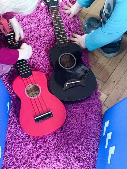 Na różowym dywanie leżą ukulele. Dzieci podejmują je z podłogi.