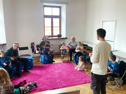 Dzieci z rodzicami siedzą w półkole i grają na ukulele.