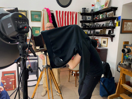 Kobieta ma narzuconą na głowę bluzę. Próbuje zrobić zdjęcie aparatem sprzed 100 lat.