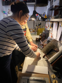 Kobieta i fotograf przebywają w ciemni fotograficznej. Kobieta rysuje coś na mokrym materiale. 