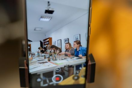 Uczestniczki warsztatów siedzą przy stole i malują. Widać je na ekranie telefonu. Ktoś chce im zrobić zdjęcie.