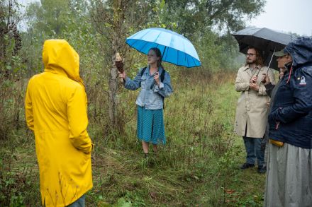Kobieta trzyma rybę z wikliny i opowiada o swojej pracy. Wszyscy stoją na skraju lasu.