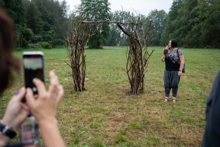 Kobieta trzyma w dłoniach telefon i robi zdjęcie innej warsztatowiczce. Stoi ona przy swojej pracy. To łuk z wikliny.