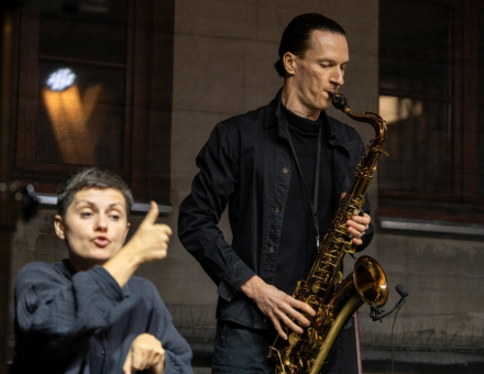 Mężczyzna gra na saksofonie. Obok niego kobieta miga.