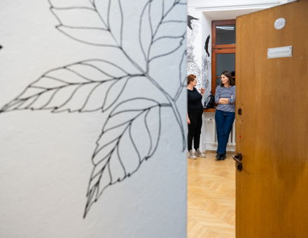 Otwarte drzwi prowadzą do sali wystawowej. Są w niej dwie kobiety. Otwarte drzwi prowadzą do sali wystawowej. Są w niej dwie kobiety.