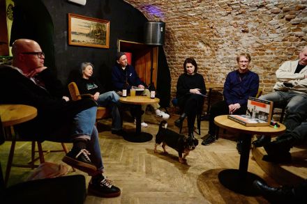 Uczestnicy spotkania siedzą przy stolikach barowych i słuchają.