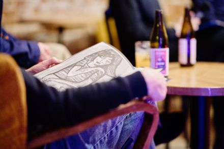 Na kolanach mężczyzny leży książka z zinami. Na stoliku przed nim stoi butelka piwa.