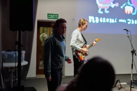Mężczyzna gra na gitarze. Obok stoi kobieta i gestykuluje. 