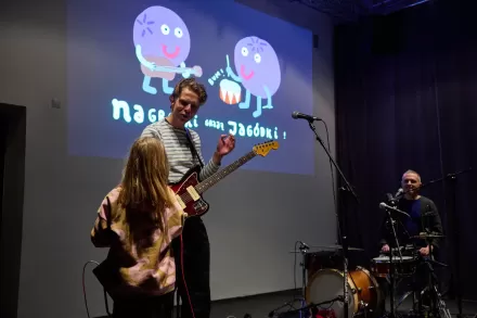 Dwaj mężczyźni grają na gitarze i perkusji. Obok nich śpiewa dziewczynka