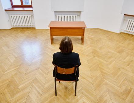 Na krześle siedzi kobieta. Widzimy ją tyłem.  Przed nią stoi biurko.