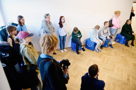 Grupa dorosłych i dzieci siedzi dookoła sali i słuchają. 