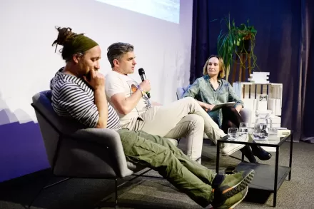 Przy stoliku na kanapie siedzą dwaj mężczyźni i kobieta. Rozmawiają,