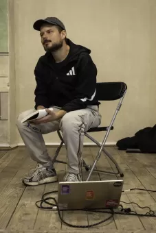 Prowadzący siedzi na krześle, a na podłodze sto laptop. 