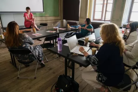 Uczestniczki wydarzenia słuchają prowadzącej. Siedzą przy stole, a prowadząca na blacie. 