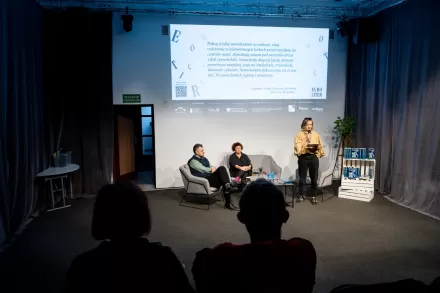 Na fotelach przy stoliku siedzą kobieta i mężczyzna. Obok stoi kobieta i przemawia.