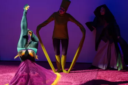 Trójka aktorów chodzi po różowym dywanie. Światło jest przyciemnione. 