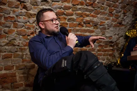 Prowadzący siedzi a krześle, mówi do mikrofonu i gestykuluje. 