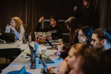 Uczestnicy siedzą przy stolikach. Mają rozłożone zeszyty i laptopy. Jeden chłopak podnosi rękę.