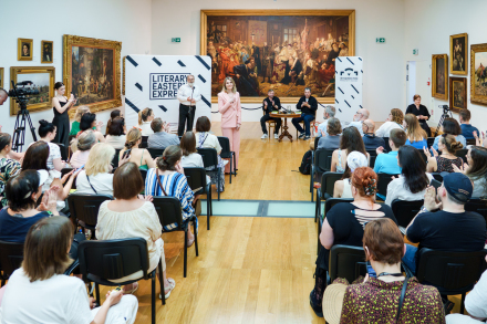 Cała sala jest pełna publiczności. Ludzie siedzą na krzesłach lub stoją.