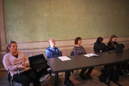 Uczestnicy spotkania siedzą przy stole i słuchają wystąpienia.