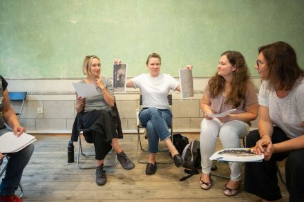 Kobieta trzyma w rękach skany dwóch zdjęć i kieruje je w stronę uczestniczek warsztatów. Jej ręce są skierowane w prawą i lewą stronę.