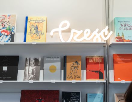 Zdjęcie dwóch regałów z ustawionymi książkami oraz świecącym napisem ,,Cześć".