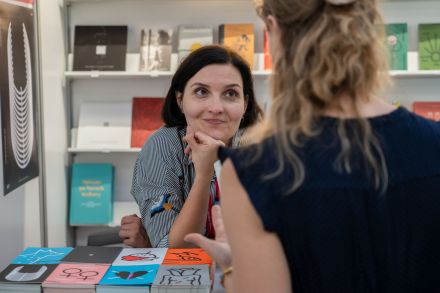 Kobieta oparta o blat. na którym są rozłożone książki, rozmawia z drugą kobietą odwróconą  plecami do aparatu.