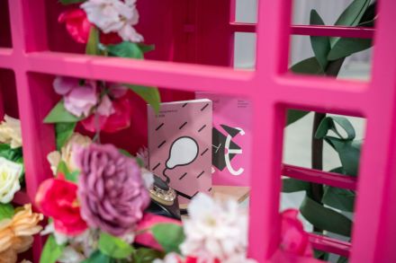 Zdjęcie jasno różowej książki z białym balonem oraz różowej książki z geometrycznymi kształtami, ustawionych we wnętrzu różowej budki telefonicznej ozdobionej kwiatami.