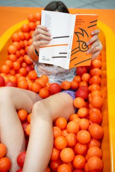 Dziewczyna siedzi w basenie z pomarańczowymi kulkami i trzyma przed sobą książkę z biało-pomarańczową okładką , która zasłania jej twarz.