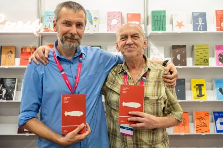 Zdjęcie dwóch mężczyzn, obejmujących się rękami, trzymających dwie czerwone książki z białym pociskiem,, na tle regałów z książkami. 