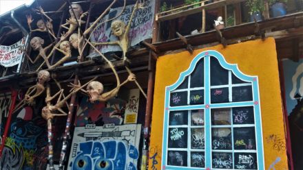 Chude stworki zawieszone na poręczach i rurkach budynku o różnych wyrazach twarzy. Budynek jest cały pokryty graffiti oraz banerami. W oczy rzuca się  żółta ściana z oknem z niebieską ramą, podzielonym na małe okienka, w których są napisy. 