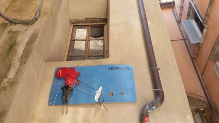 Niebieska tablica zawieszona na ścianie budynku pod oknem, przedstawiająca czarnego ludzika wspinającego się na kontynent Afryki, a poprawej stronie biały ludzik próbuje dostać się tam na linach. Obok białego ludzika po prawej stronie są dwie nieregularne dziury. W prawym górnym rogu widnieje napis ,,JEPETTU".