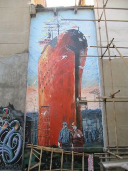 Mural przedstawia dwóch mężczyzn, którzy stoją na tle bardzo wysokiego i ogromnego, czerwonego statku.
