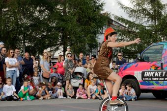 Plenerowy pokaz sztuki cyrkowej, na pierwszym planie artysta jadący na monocyklu,  w tle zgromadzona publiczność.