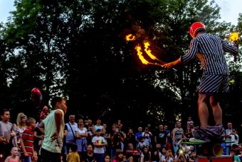 Plenerowy pokaz sztuki cyrkowej,  zdjęcie ukazuje artystę  utrzymującego równowagę  na rola boli, trzymając płonące pochodnie w dłoniach, w tle publiczność.