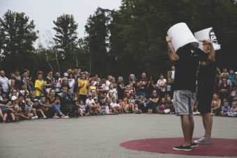 Plenerowy pokaz sztuki cyrkowej, dwóch artystów stojących na środku z wiadrami na głowach, w tle publiczność.