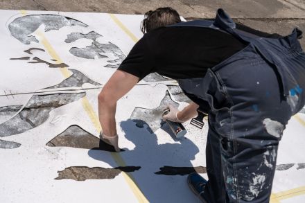Mężczyzna, białym sprayem, odrysowuje mural od ułożonego szablonu na zboczu.