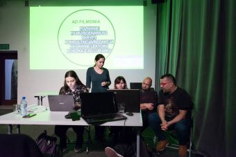 Na zdjęciu uczestnicy pracują w grupach, siedząc przed komputerami, za nimi wyświetlony jest slajd ( Ad filmowa).
