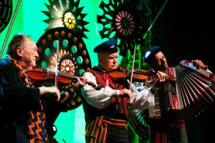 Trzej muzycy w strojach ludowych dają koncert na scenie. Dwóch gra na skrzypcach, a jeden na akordeonie.