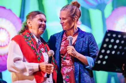 Scena. Kobieta w niebieskiej marynarce z mikrofonem, obejmuje ramieniem starszą kobietę ubraną w strój ludowy, również trzymającą mikrofon.