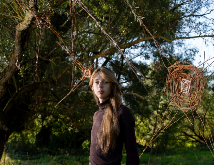 Dziewczyna stoi koło swojej pracy. To pajęczyna z wikliny zwisająca z drzewa.