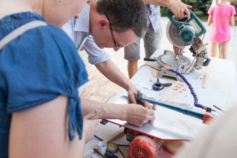 Na zdjęciu uczestnicy warsztatu obrabiają, tną drewniane elementy przy użyciu maszyn.