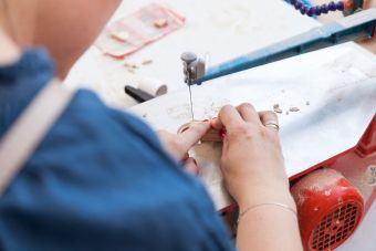 Plenerowy warsztat w ramach lekcji sztuki. Zbliżenie na  damskie dłonie. Kobieta trzyma drewniany element i docina go na maszynie do obróbki drewna.